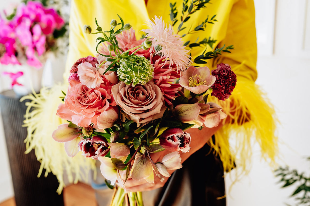 Full hand-held bouquet of peach and pink roses, burgundy blooms and greenery.